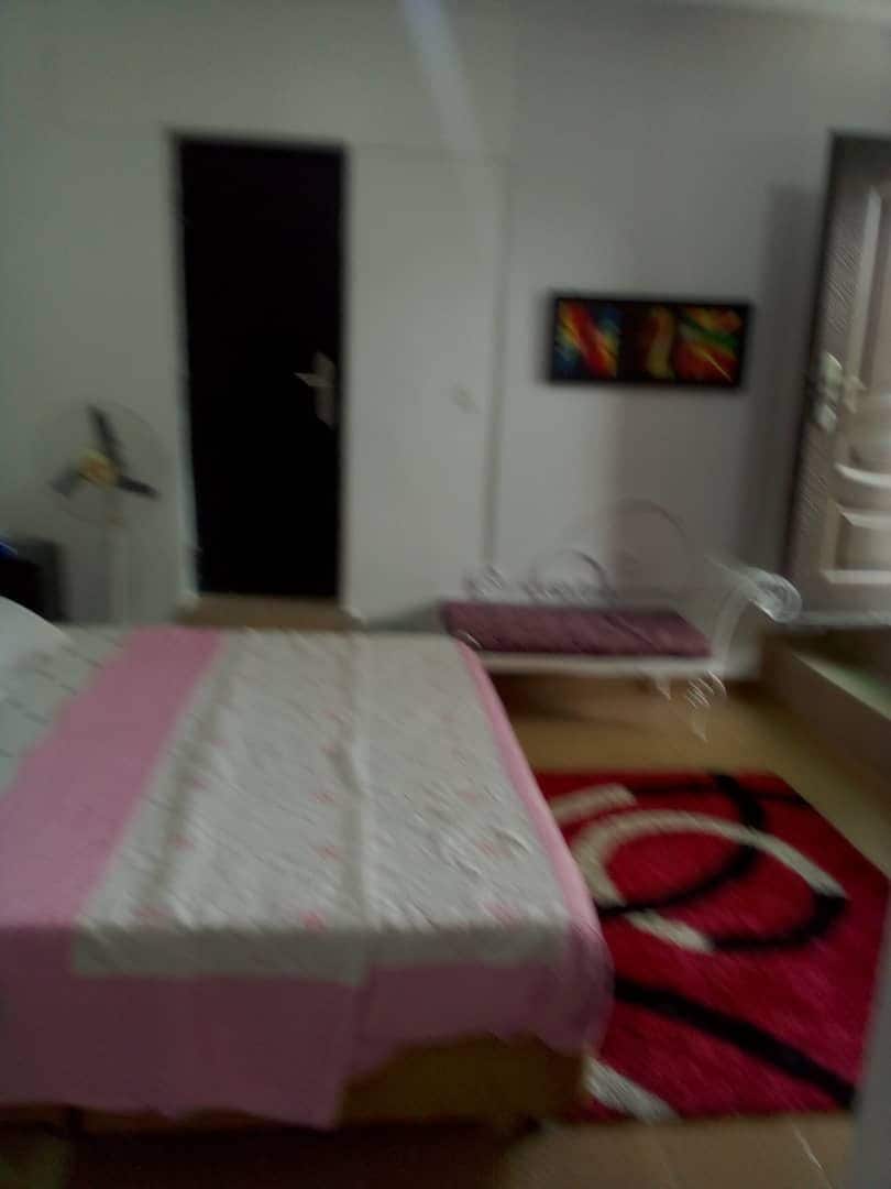 A cozy sleeping area features a bed with a patterned cover and a prominent rug beneath. A colorful wall art piece adds a touch of vibrancy, while a fan is observed near the bed and a door leads to another space.