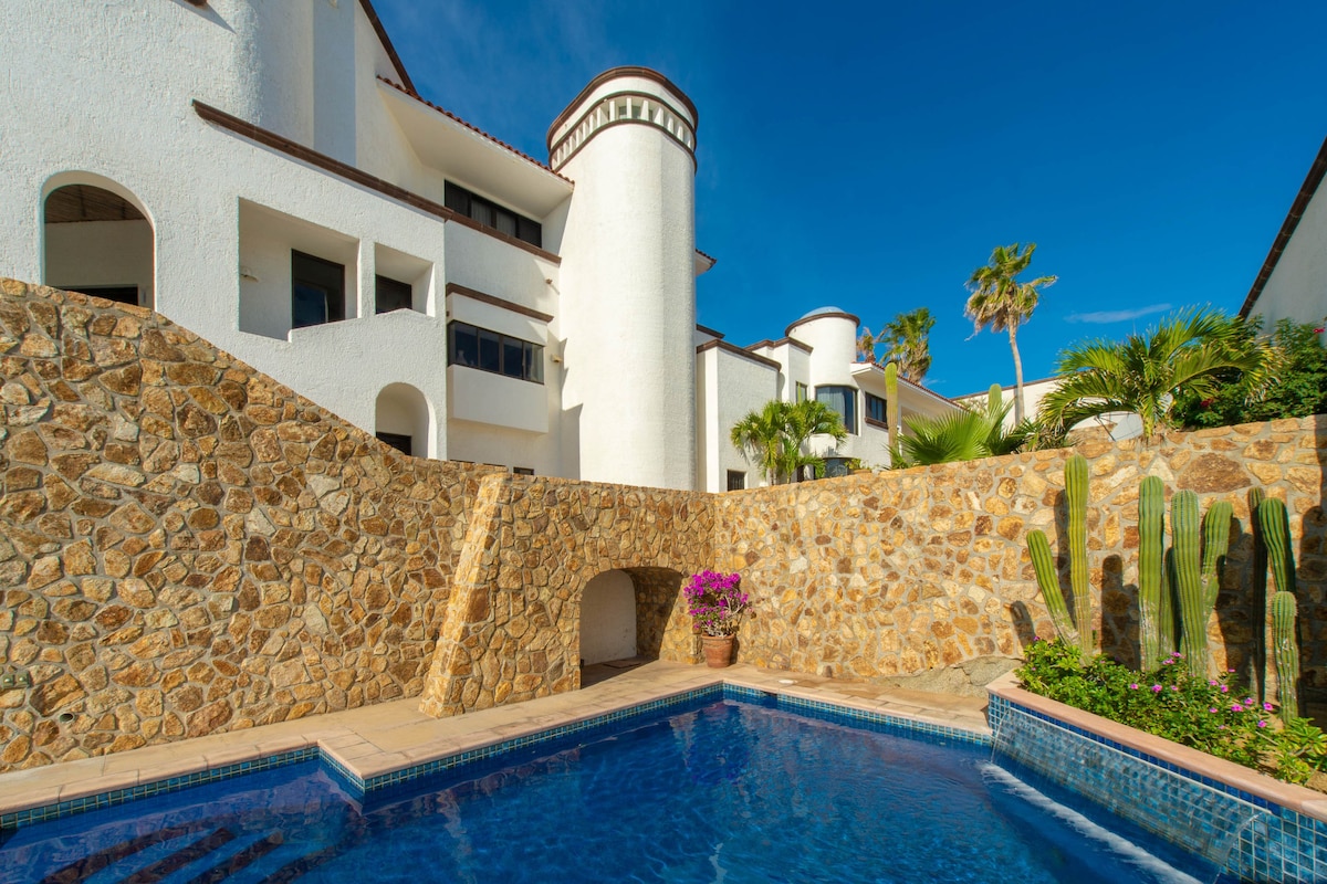 A sunlit area features a stone wall surrounding a pool, accented by vibrant tropical plants. The architecture includes white walls and distinct tower structures, set against a clear blue sky, providing an inviting outdoor space for relaxation.
