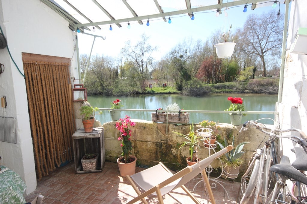 A terrace overlooks the Charente River, surrounded by greenery. Potted plants, including vibrant flowers, add color to the space. A small wooden table is present, along with a folding chair and bicycles are leaned against the wall, enhancing the outdoor setting.