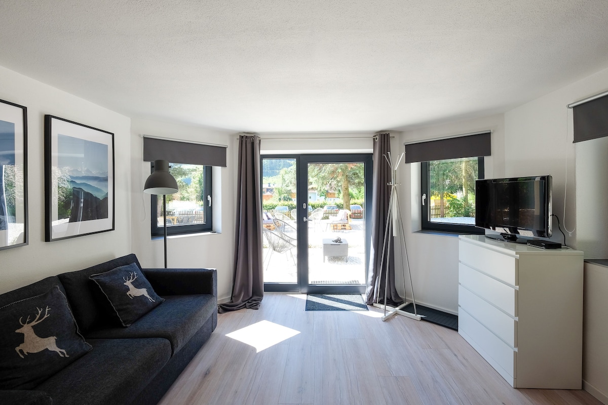 A cozy living area is illuminated by natural light from large windows. A dark sofa with decorative cushions complements the light wood flooring. A flat-screen TV and a dresser are positioned nearby, while outdoor views are framed by gray curtains.