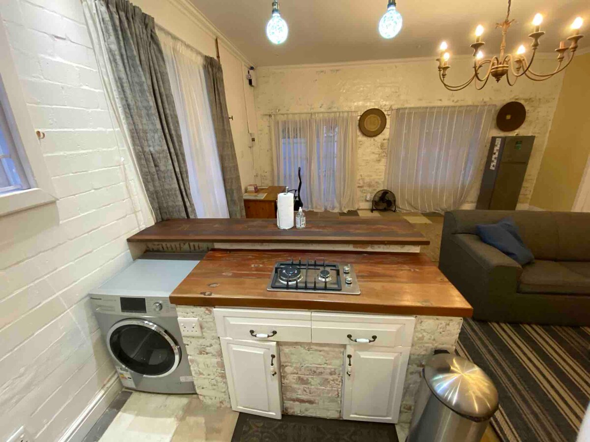 A functional kitchen area is presented with a wooden countertop and a gas stove. A washing machine is positioned beside the counter, and a trash bin is located below. Soft lighting is provided by pendant fixtures, with a chandelier adding an elegant touch to the room.