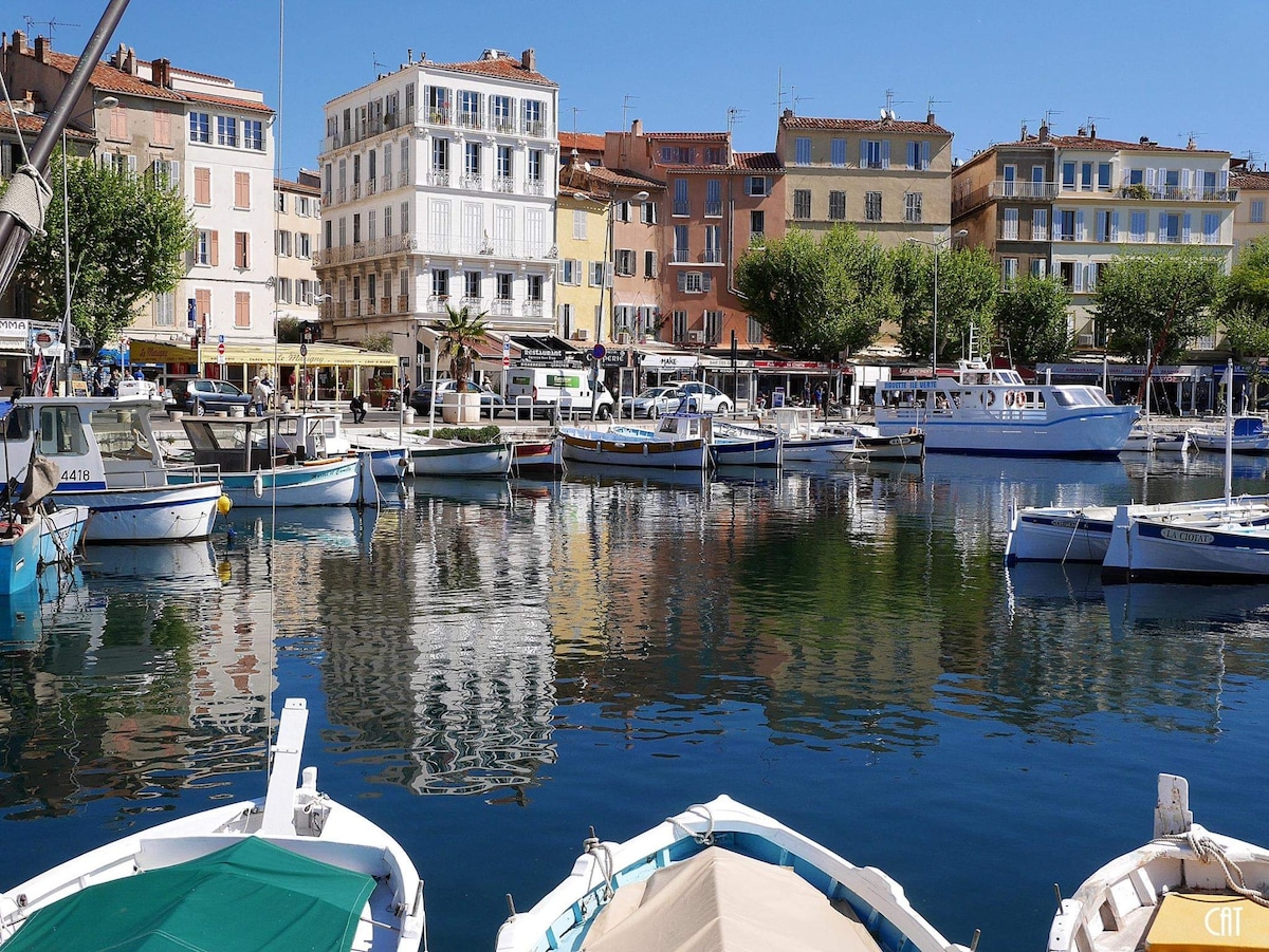 A marina scene is depicted with several boats docked in calm water, reflecting the vibrant colors of surrounding buildings. The three-story structures feature shutters and balconies, while greenery from nearby trees frames the waterfront, creating a relaxed atmosphere.