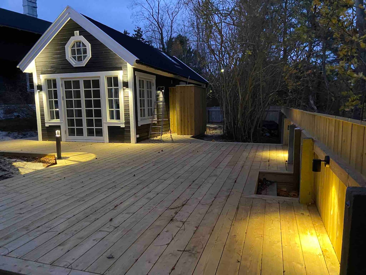 The outdoor area is illuminated by soft lighting, highlighting the spacious wooden deck that surrounds the guesthouse. Large glass doors lead into the interiors, while the surrounding trees add an element of privacy to the setting.