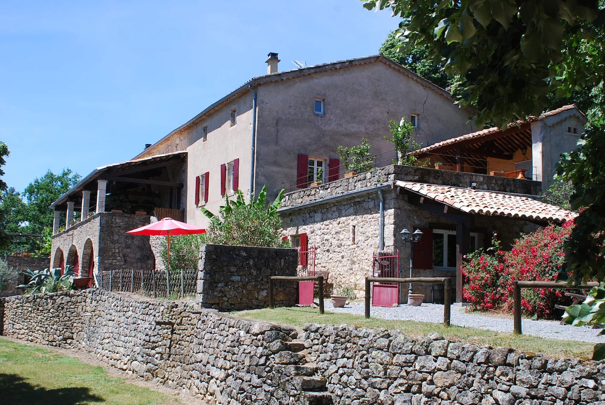 The exterior of a rustic stone house is captured, featuring pink-shuttered windows and a textured façade. A shaded terrace is visible with dining furniture and a red umbrella, while lush greenery and flowering plants enhance the surrounding landscape. A stone wall separates the house from a gravel path.