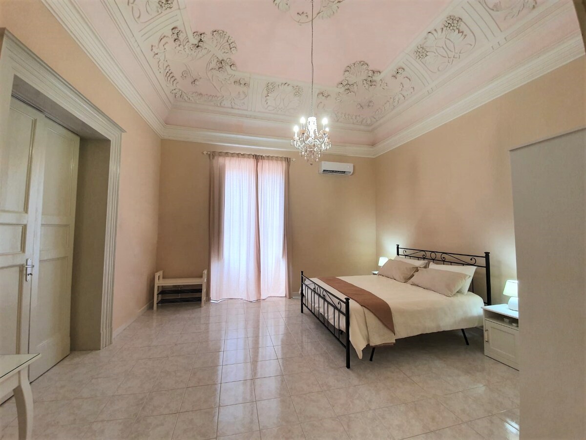 A spacious room features a large bed with soft bedding. A delicate chandelier hangs from a beautifully adorned ceiling. Natural light filters through a tall window with sheer curtains, illuminating the light-colored walls. A small seating area and nightstands are also visible.