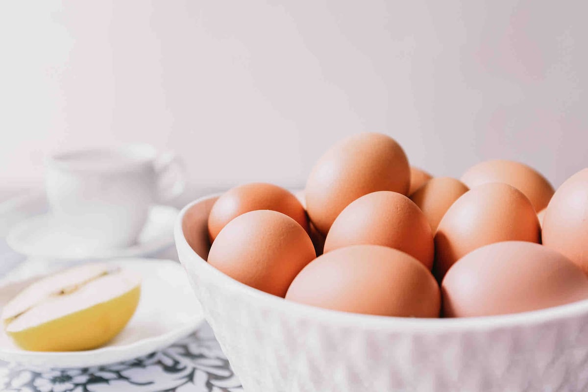 A bowl filled with numerous brown eggs is positioned prominently in the foreground. A small plate featuring a sliced pear is placed nearby, along with a cup and saucer set on a patterned tablecloth.
