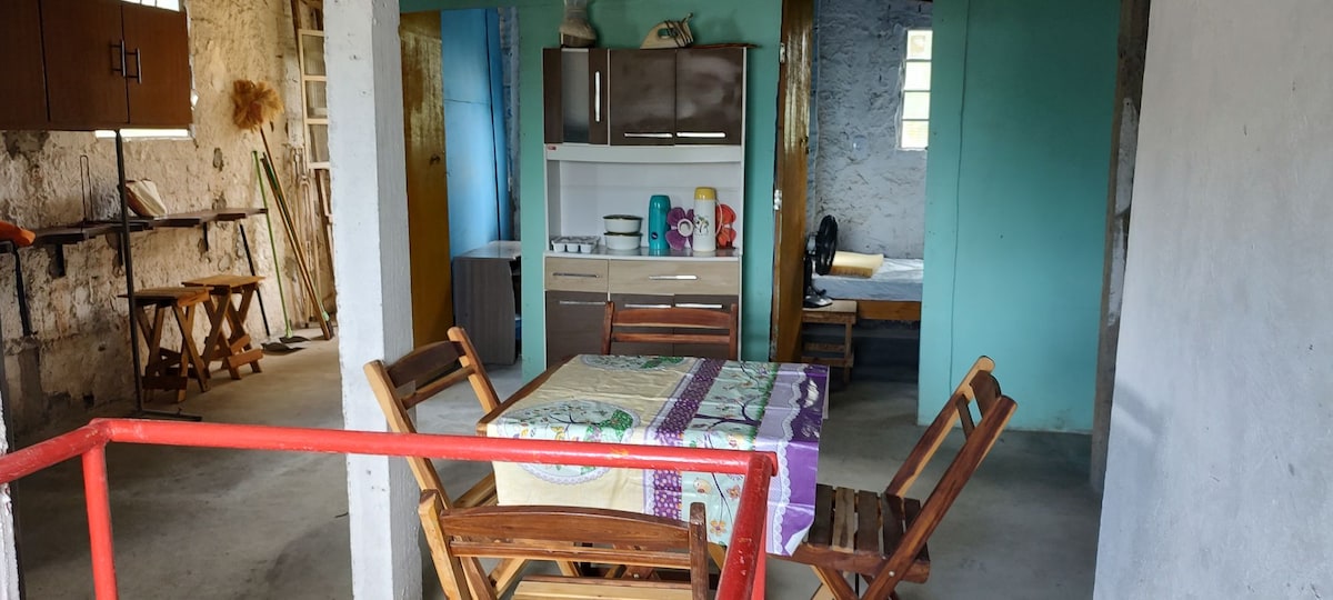 A spacious area features a rectangular dining table surrounded by wooden chairs. A kitchen with cabinetry and appliances is visible in the background. Light enters through small windows, illuminating both the dining space and a sleeping area in the distance.