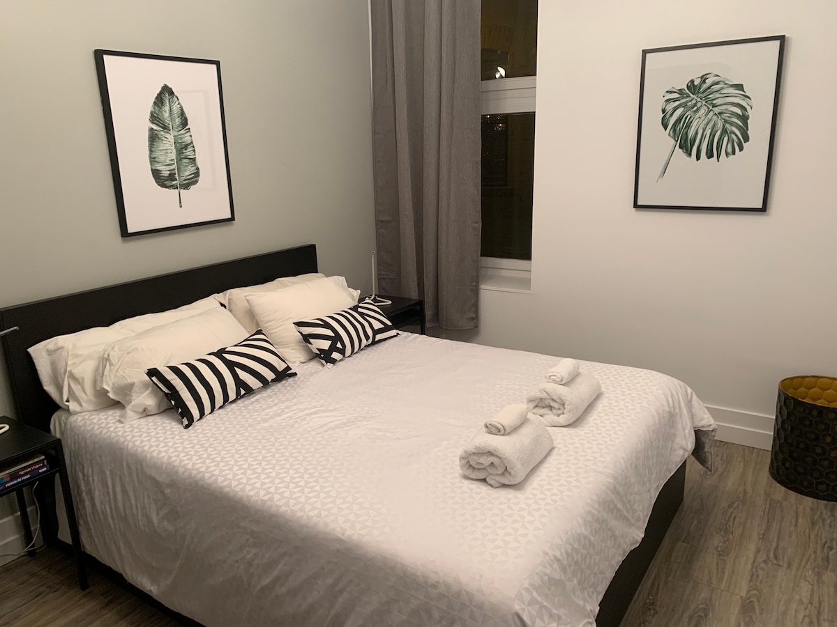 A cozy sleeping area features a spacious bed adorned with white linens and decorative striped pillows. Two framed botanical prints are displayed on the walls. Neatly rolled towels are arranged at the foot of the bed, and a small accent table is visible beside it.