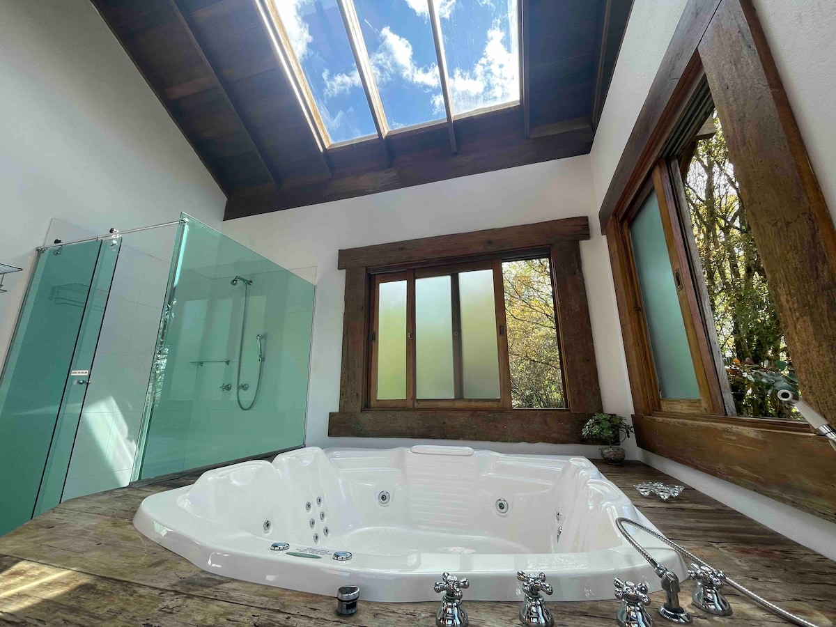 A private relaxation space features a spacious hot tub surrounded by wooden accents and natural light. A skylight above provides a view of the sky, complementing large windows that frame the serene outdoor landscape. A sleek glass shower enclosure is visible in the corner.