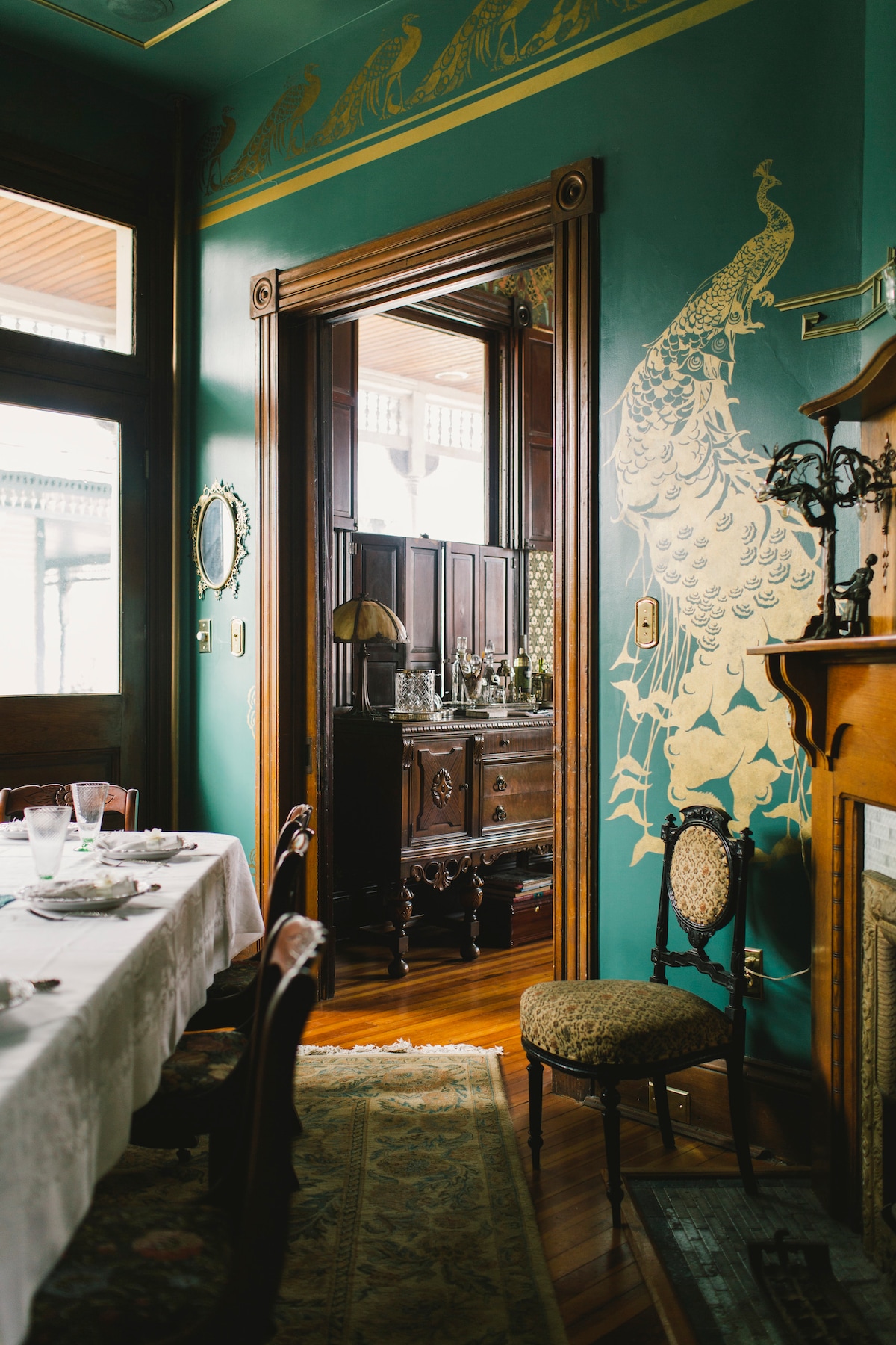 A dining area features a long table set with dishes and glassware, surrounded by ornate chairs. The walls are painted in a rich teal adorned with golden peacock motifs. An elegant wooden sideboard is visible in the adjoining room, complemented by period detailing and wooden flooring.