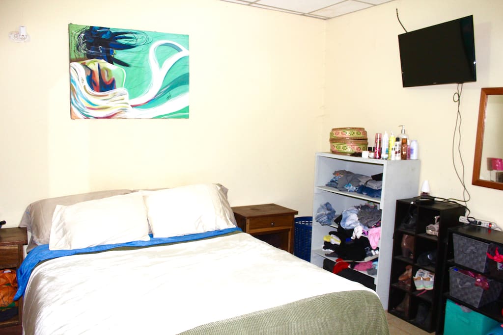 Bedroom with shelves, closet space, flatscreen TV, AC and bathroom access. Shelves are cleaned out for guests. Bedside tables have extension cords and outlets for charging devices. Bed has soft sheets and extra pillows.