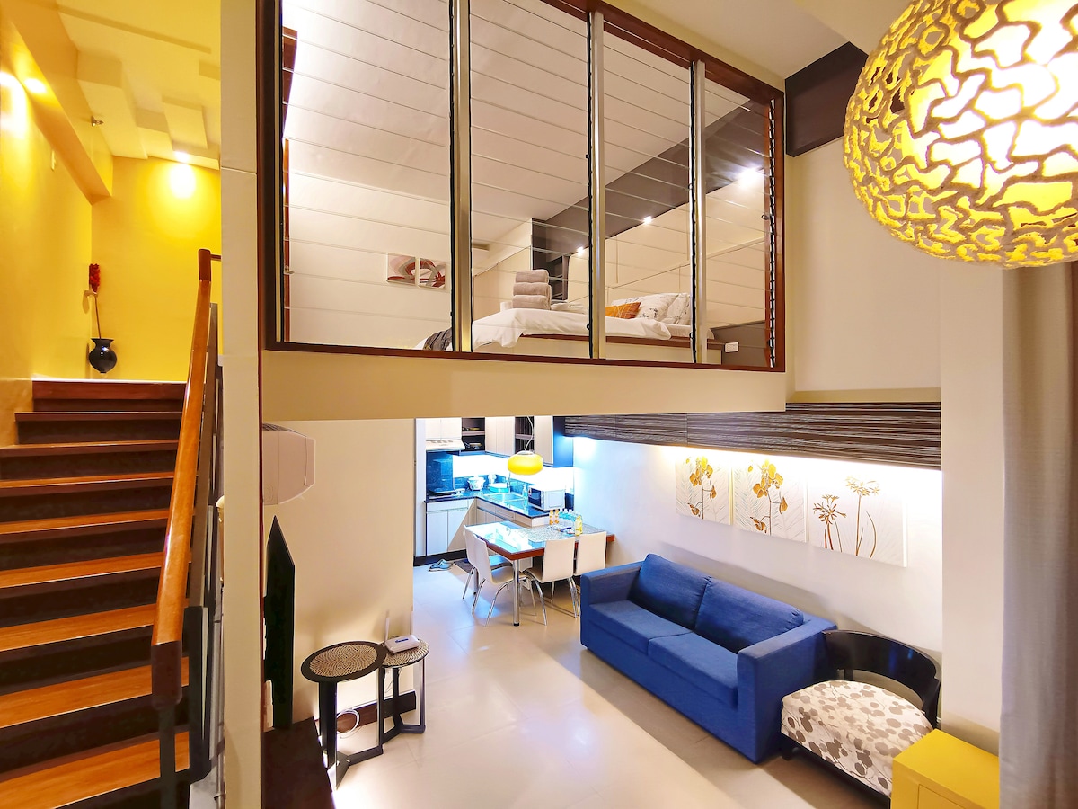 A view of the loft-style living area showcases a spacious layout with a blue sofa, dining table, and well-equipped kitchen. Stairs lead to an upper sleeping area, visible through glass railings, where a bed is neatly arranged. Natural light fills the room, enhancing the modern decor.