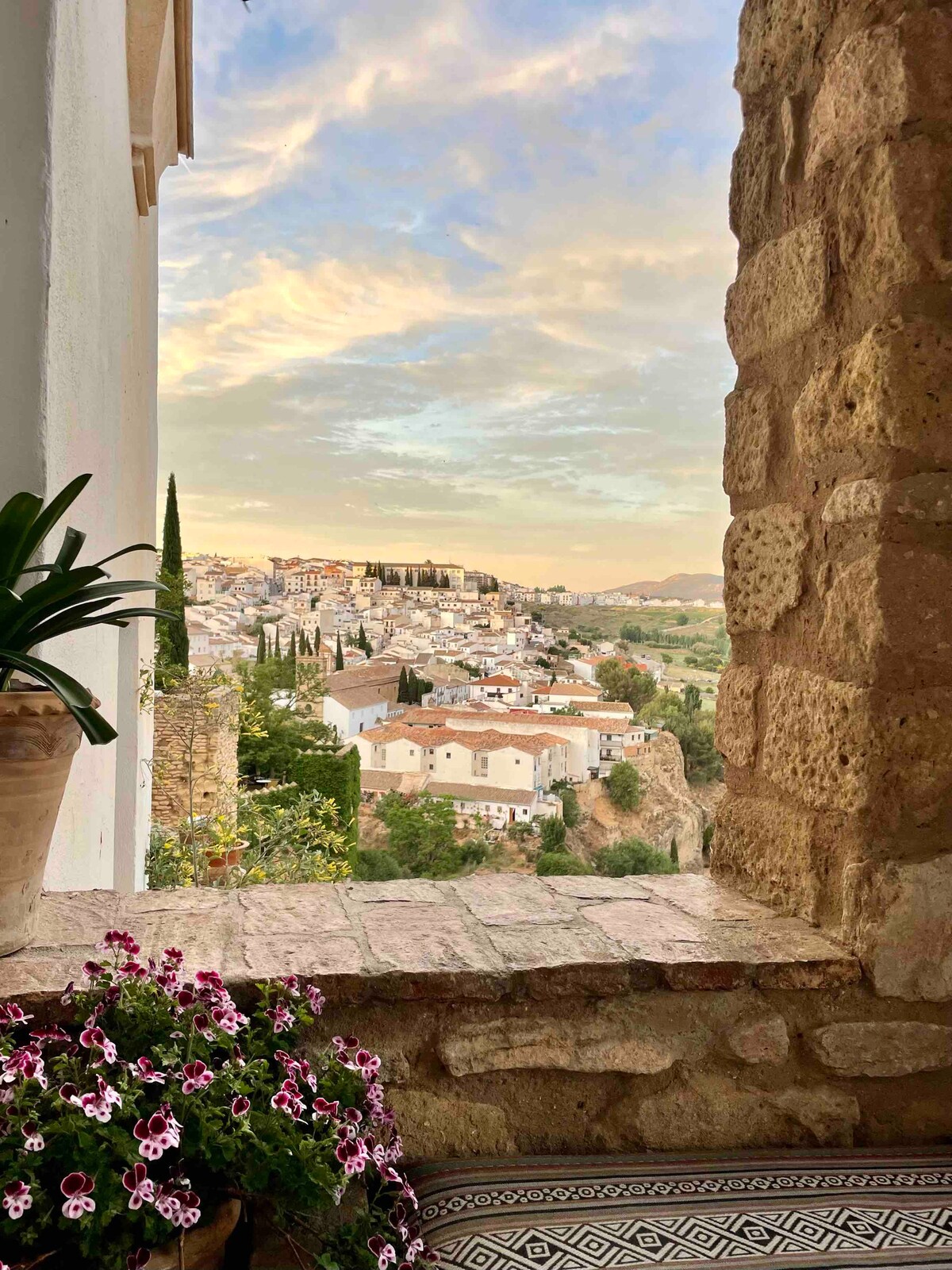 An arched stone window frames a panoramic view of the historic city, showcasing charming white buildings nestled among lush greenery. A decorative pot with vibrant flowers brightens the foreground, while the sky exhibits soft pastel hues during sunset.