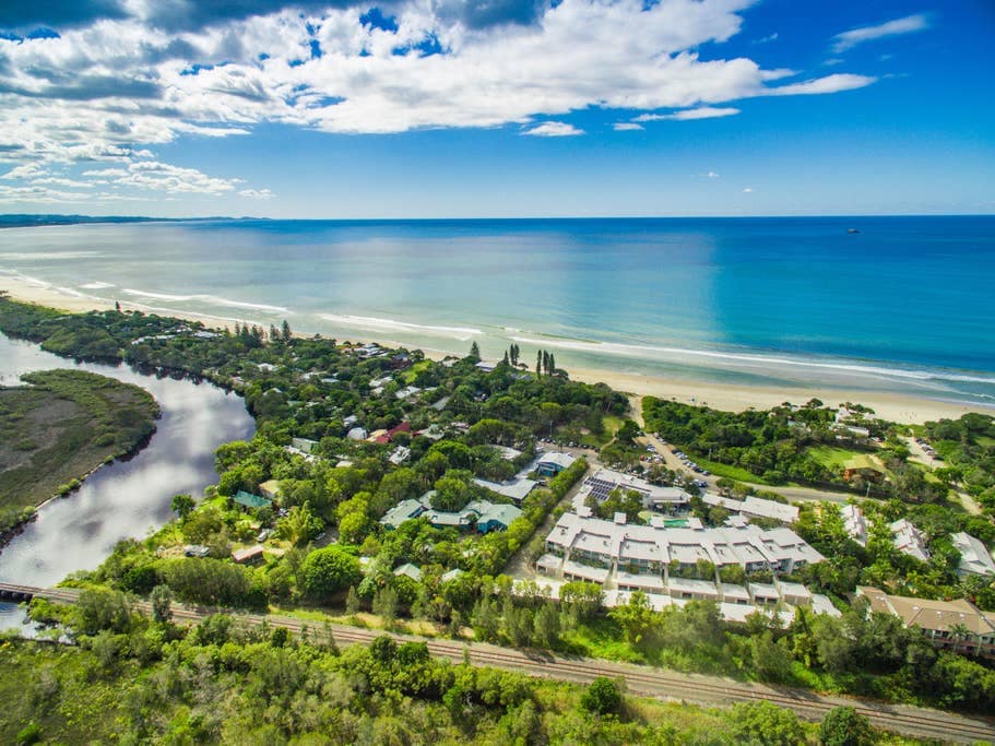 An aerial view captures the coastline alongside lush greenery, revealing the tranquil beach and sparkling ocean. The image showcases both the residential area and the water bodies, with gentle waves lapping at the shore under a clear blue sky.