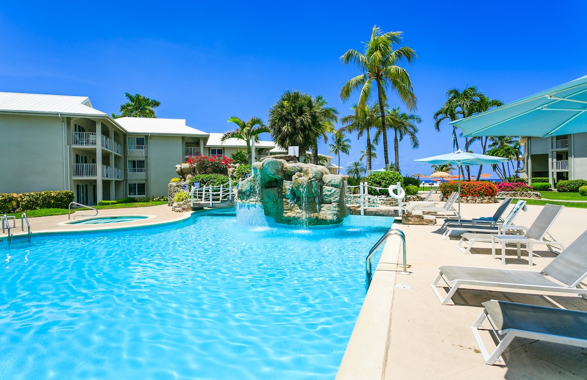 A sparkling pool is framed by tropical palm trees and vibrant flowering plants, creating a serene outdoor setting. Lounge chairs are arranged around the pool area, inviting relaxation. The backdrop features a two-story building, with expansive balconies and access to the lush grounds.