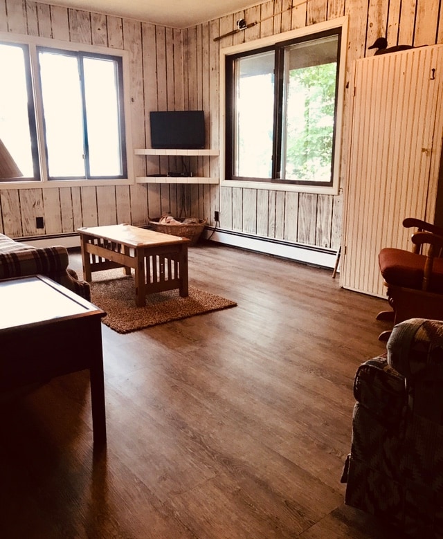 A spacious living area is characterized by wooden panel walls and large windows that invite natural light. A coffee table sits centered on a textured rug, with a comfortable couch and chair providing seating options. A small television and shelves are visible along one wall.