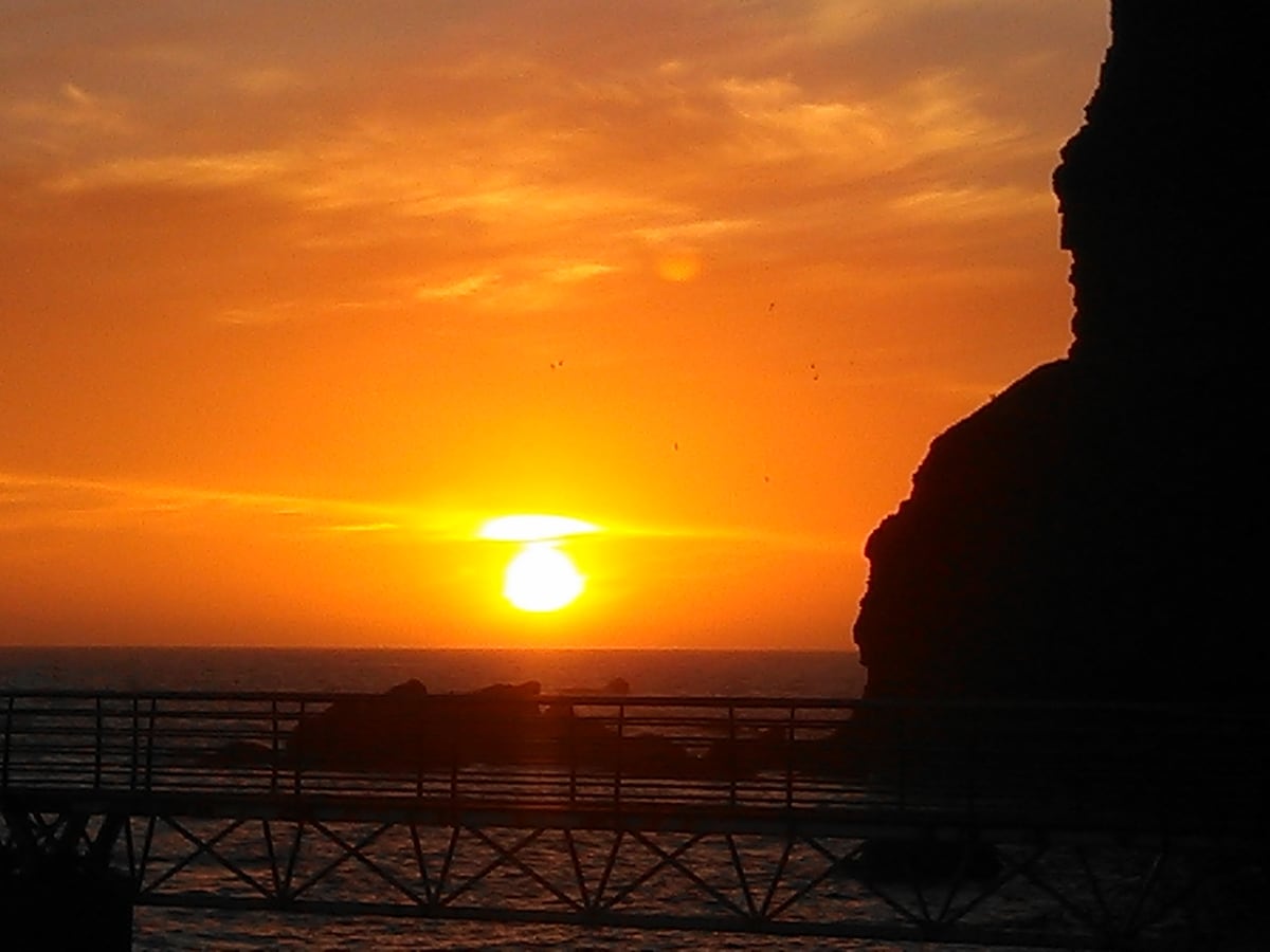A vibrant sunset casts orange and yellow hues across the sky, reflecting on the ocean's surface. Silhouettes of rocky cliffs frame the scene, while a bridge is visible in the foreground, leading towards the water.