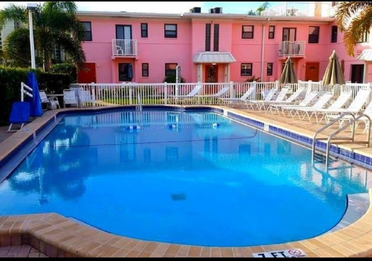 A clear blue pool sits surrounded by lounge chairs under sunny skies. The area is bordered by a white fence, and pink buildings provide a vibrant backdrop. Several umbrellas are positioned over tables nearby, enhancing the recreational atmosphere of the resort.