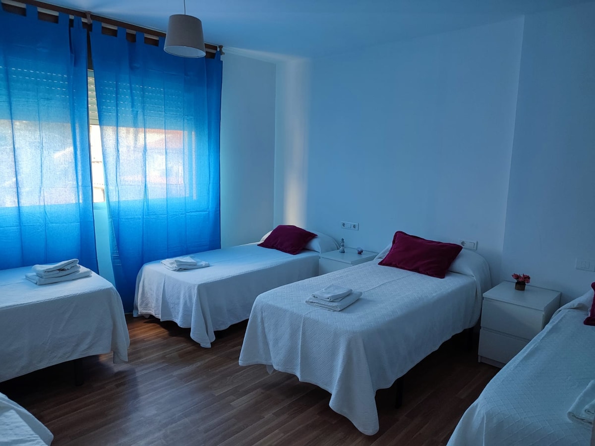 A bright room features four spacious beds, each neatly made with white linens. Vibrant blue curtains allow natural light to filter in, enhancing the clean and fresh atmosphere. Two nightstands are positioned beside the beds, providing additional space for personal items.