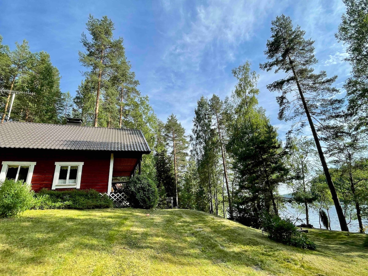 A Finnish kota is positioned among tall trees on a green lawn, with a clear view of a nearby lake. The building is characterized by its red wooden exterior and a sloped roof, blending harmoniously with the natural surroundings under a blue sky.