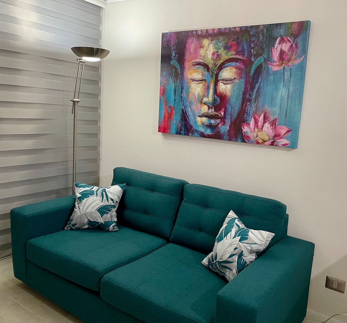 A modern living area features a teal sofa adorned with two decorative pillows. Above the sofa, a colorful artwork of a serene Buddha face is displayed, complemented by abstract floral elements.