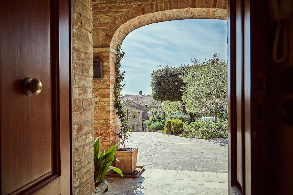 The view from the doorway captures a charming garden area, framed by rustic stone walls. Lush plants are visible on either side, while cobblestone paths lead towards well-kept greenery and distant buildings, under a clear sky.