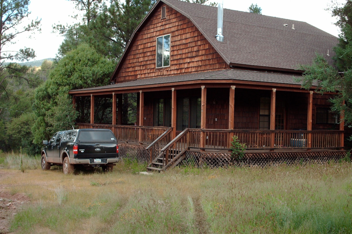 A two-story cabin with wooden shingle siding is surrounded by greenery. The spacious wrap-around porch features a railing and multiple entry points. A parked dark vehicle is visible in front of the cabin, set against a natural landscape of grass and trees.