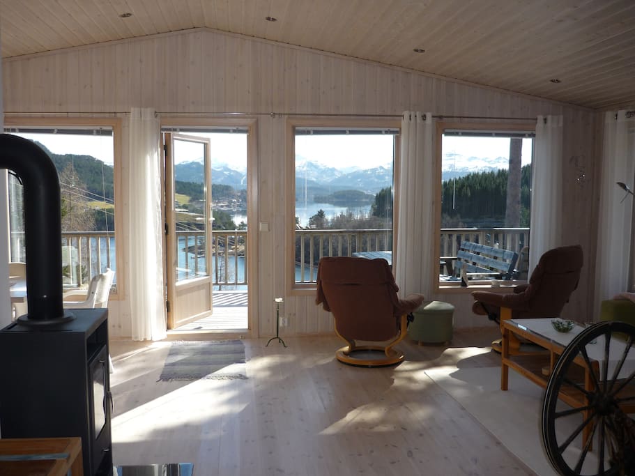 Stua med 6 store vinduer mot sjøen og verandadør.
Vedovn og sittegruppe.