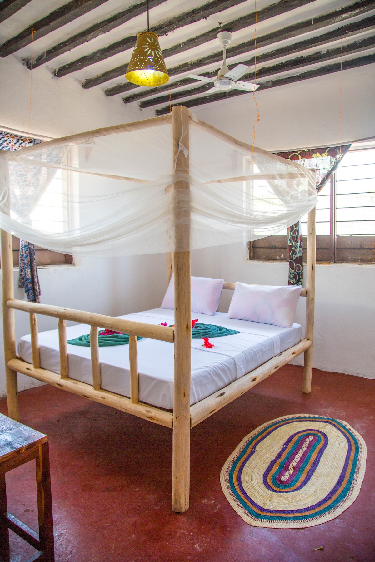 A simple bedroom features a four-poster bed with a light canopy, adorned with decorative pillows. Natural light filters through window screens, and a woven rug adds warmth to the terracotta floor. A wooden table sits nearby, completing the cozy atmosphere.