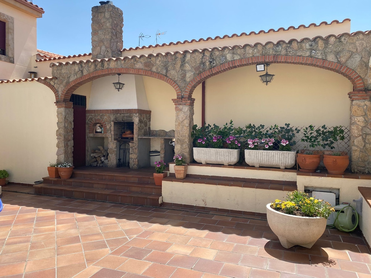 An outdoor patio area is featured, complete with stone arches and a built-in barbecue. Potted plants with colorful flowers are positioned along the wall, adding natural touches to the setting. The space is paved with tiles, enhancing its rustic charm.
