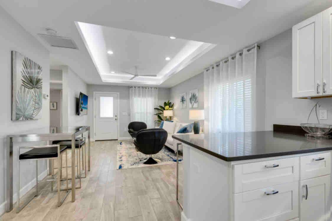 A modern open-concept living space features stylish furnishings and natural light from large windows. A comfortable seating area includes black chairs and a light-colored sofa, complemented by artistic décor. A sleek kitchen island with seating provides additional space for dining and socializing.