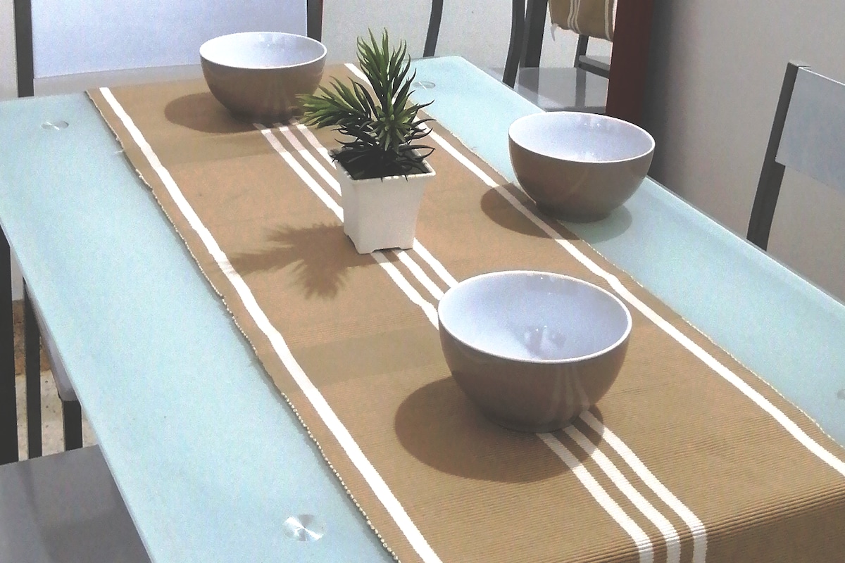 A dining table is set with a neutral-colored table runner. Three white bowls are positioned on the table, accompanied by a small potted plant that adds a touch of greenery. The glass tabletop reflects a modern design, emphasizing simplicity and functionality.