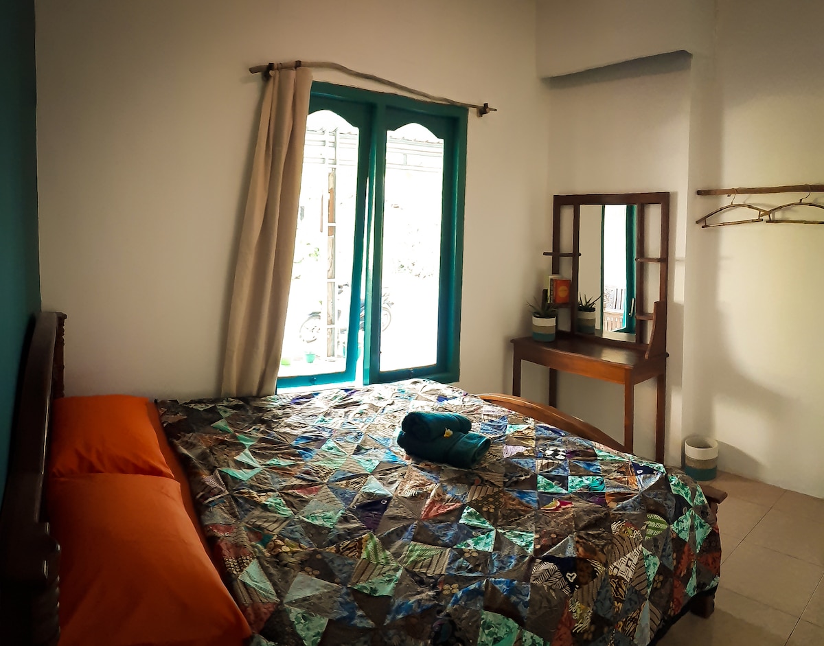 A spacious bedroom is featured, showcasing a large bed adorned with a colorful patchwork quilt and orange accent pillows. Natural light filters through two vibrant teal windows, illuminating a wooden dresser with a mirror nearby. A hanging rack is positioned against the wall for convenience.