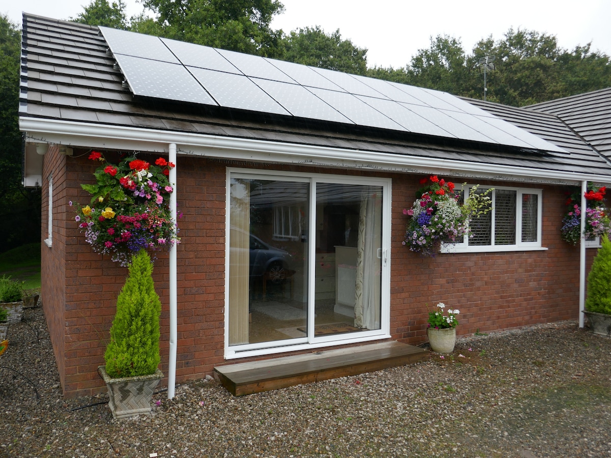The exterior of the apartment is presented, featuring a brick facade with a wooden entryway and large sliding glass doors. Flowering plants adorn the entrance, and solar panels are installed on the roof, contributing to an environmentally friendly appeal.
