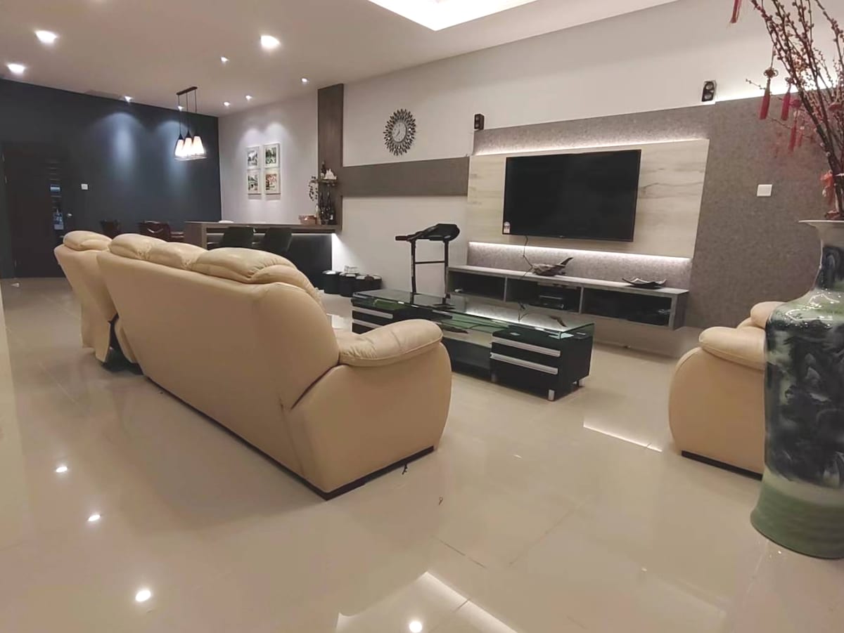 A spacious living area is presented with light-colored leather sofas facing a large mounted television. Soft lighting enhances the modern decor, while a sleek table and accent pieces create a harmonious atmosphere. The polished floor reflects the light, adding to the room's openness.