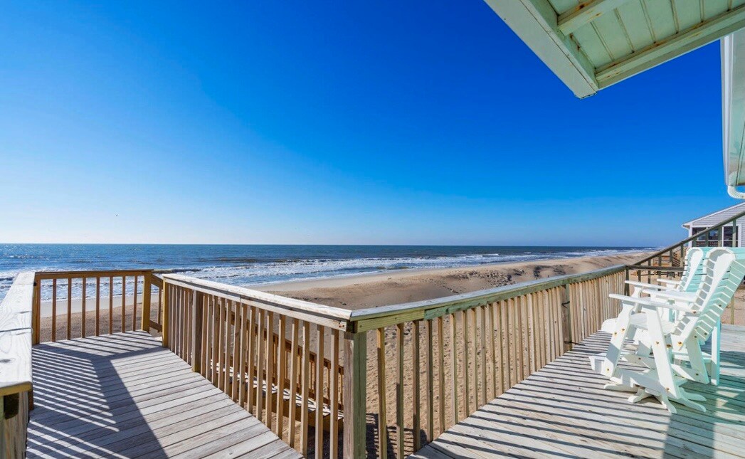 A spacious wooden deck extends towards the ocean, providing panoramic views of the sandy beach and gently rolling waves. Several white Adirondack chairs are positioned along the railing, inviting relaxation and enjoyment of the coastal scenery under a clear blue sky.
