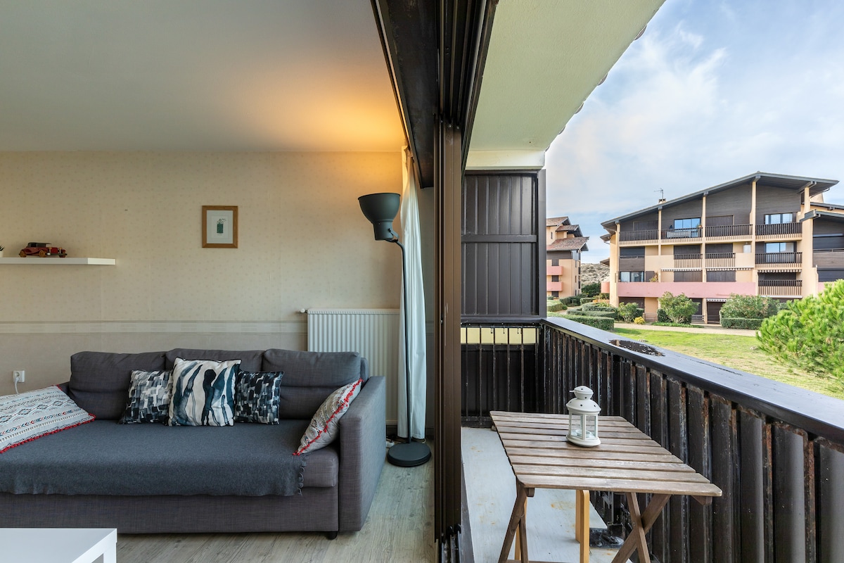 A living area features a gray sofa adorned with patterned cushions, accompanied by a small wooden table on a balcony. A lamp stands nearby, providing additional lighting. The view showcases a spacious green area and neighboring buildings beyond.