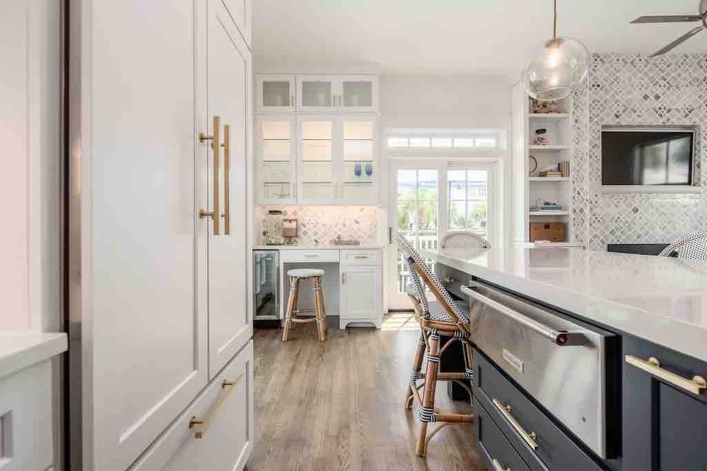 A modern kitchen is highlighted by a large island with bar seating. White cabinetry is complemented by a patterned backsplash. Natural light floods the room through a window, illuminating the elegant finishes and state-of-the-art appliances.