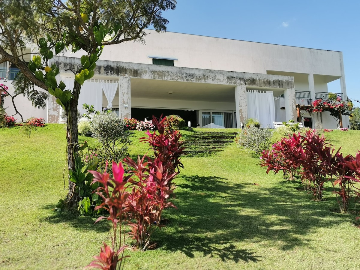 A spacious villa is positioned on a gently sloping lawn, framed by lush greenery and vibrant red plants. White drapes hang from the covered terrace, providing shade while offering views of the well-kept garden and pathway leading to the entrance.