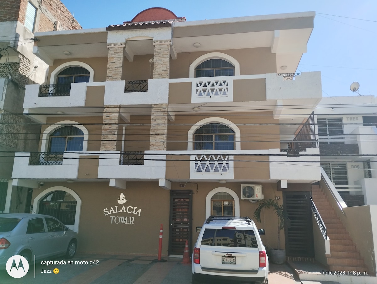 The exterior of Salacia Tower showcases a three-story building featuring a combination of stone and stucco surfaces. Balconies with decorative railings are visible on the upper floors. A parked car is situated in front of the entrance, which is framed by palm plants.