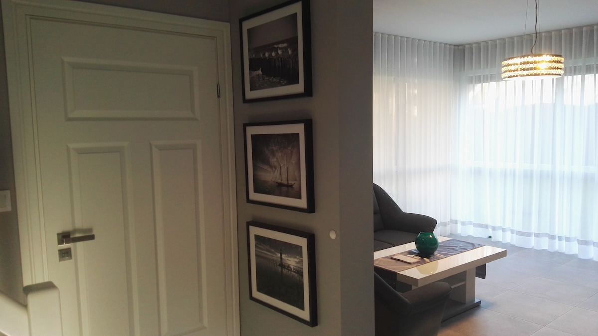 A hallway features three framed photographs, mounted on a light gray wall beside a white door. To the right, the living area is illuminated by a modern pendant light, with large sheer curtains allowing soft light to fill the space. A plush seating area is visible.