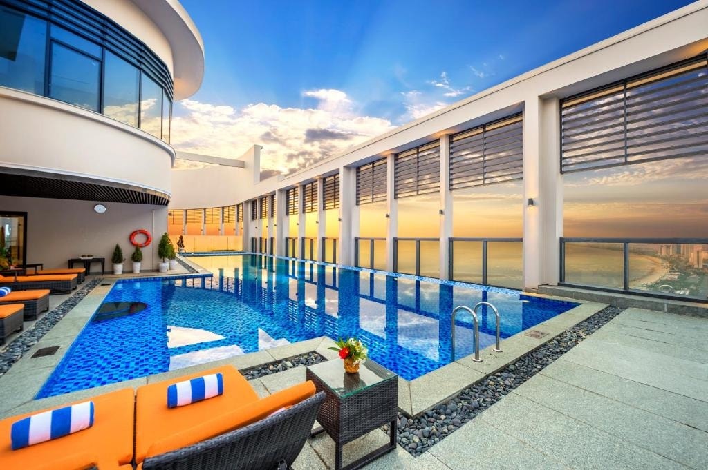 The rooftop pool area features a stunning infinity pool with vibrant blue tiles, surrounded by comfortable orange loungers. Glass panels provide an unobstructed view of the sky and coastline, while potted plants add a touch of greenery to the elegant space.