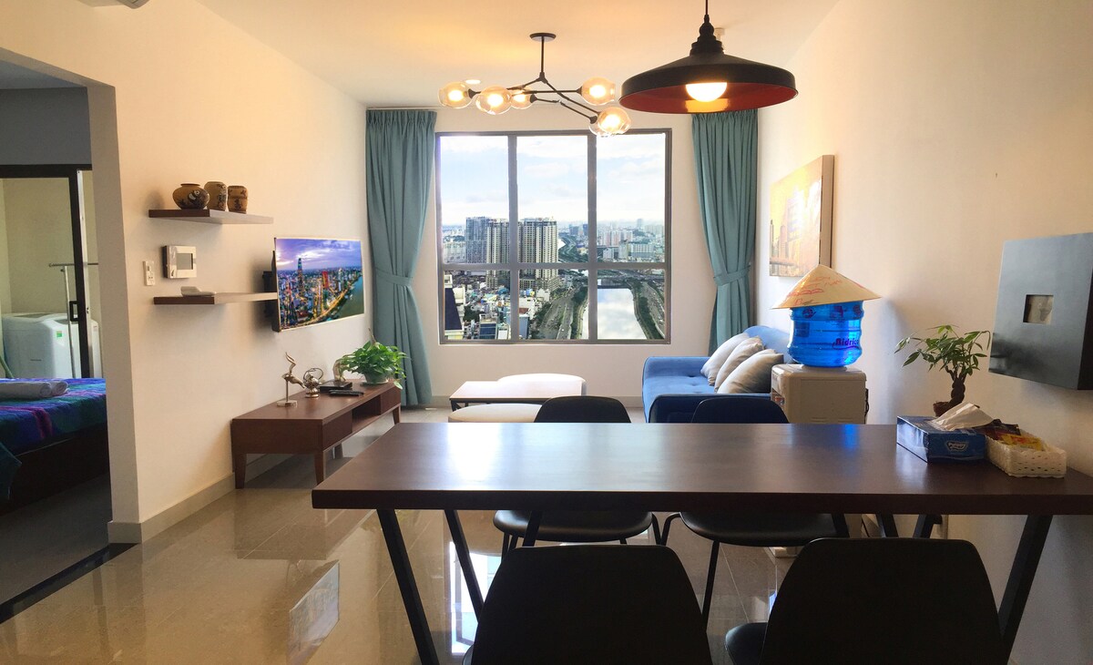A modern living area features a large window with city views, allowing natural light to fill the space. A comfortable sofa and a coffee table are arranged for relaxation, while a dining table with chairs is positioned nearby. A television is mounted on the wall, and decorative plants enhance the ambiance.