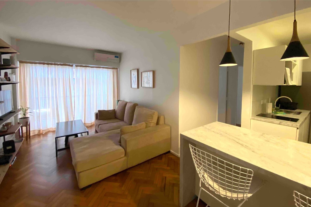 A light-filled living area features a spacious beige sectional sofa and a modern coffee table. Behind, a minimalist kitchen with a breakfast bar is complemented by sleek cabinetry and pendant lighting. Floor-to-ceiling curtains frame the large window, enhancing the bright ambiance.