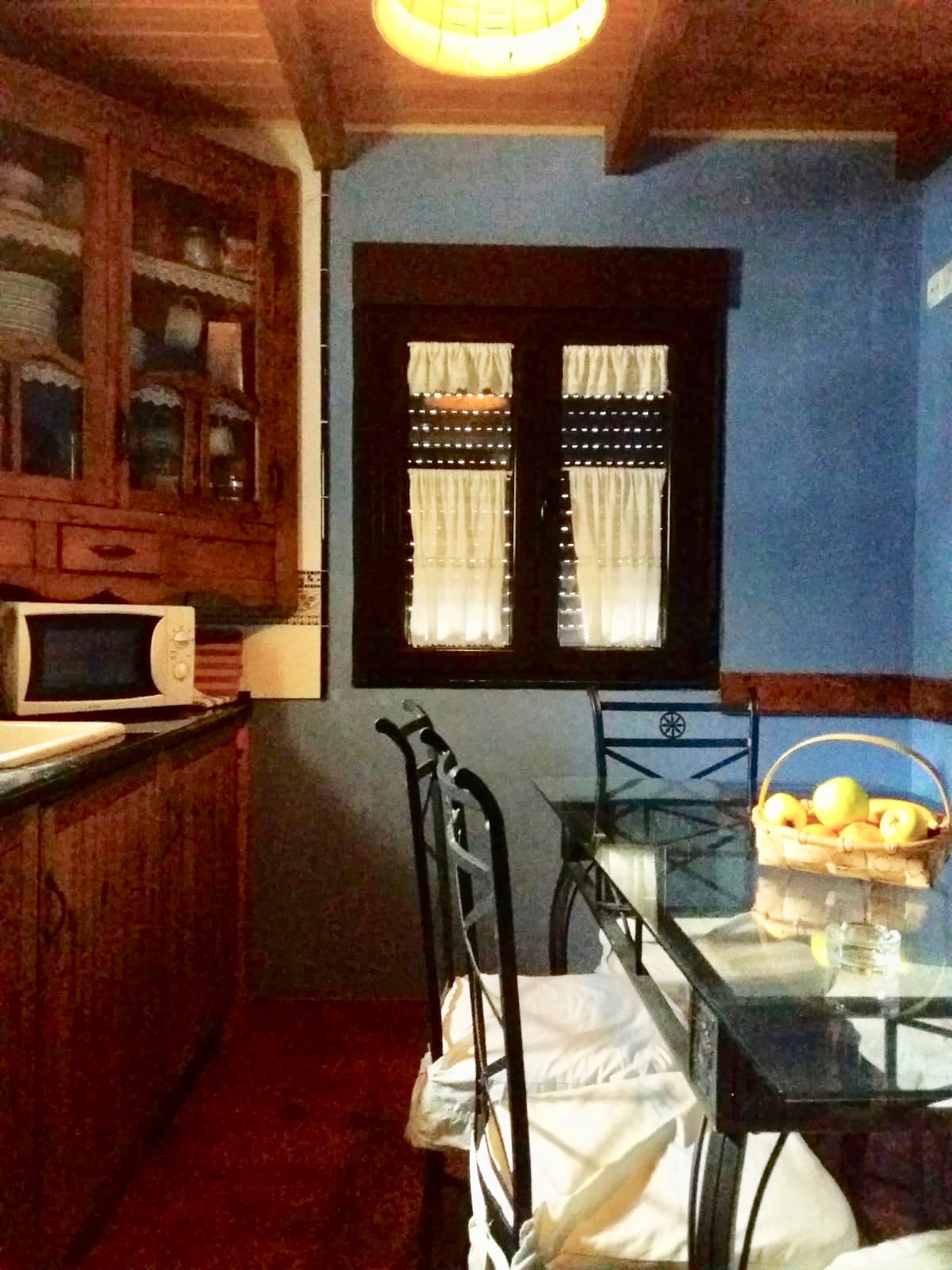 The kitchen features wooden cabinetry and a glass-top dining table accompanied by four black chairs. A microwave is visible on the countertop. Natural light enters through a window adorned with white curtains, illuminating ceramic dishware displayed in the upper cabinet.