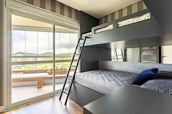 غرفة حديثة مع سرير مخطط في تشطيب مظلم، وديكور بألوان محايدة وإمكانية الوصول إلى شرفة زجاجية مع إطلالة بانورامية على المدينة والجبال. بيئة أنيقة وعملية ومضاءة بشكل جيد.