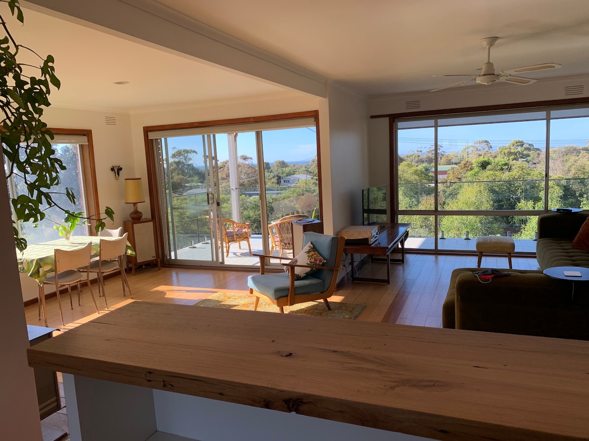An open plan living area is visible, featuring large windows that showcase views of the lush garden. A spacious sofa is positioned in the foreground, accompanied by a chair and side table. Sunlight fills the room, creating a warm environment for relaxation.