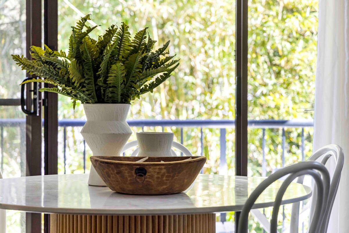 A round marble dining table is set beside large windows showcasing lush greenery. A textured white vase filled with ferns is positioned in the center, accompanied by a wooden bowl and a small white dish. Light filters through, illuminating the natural decor.