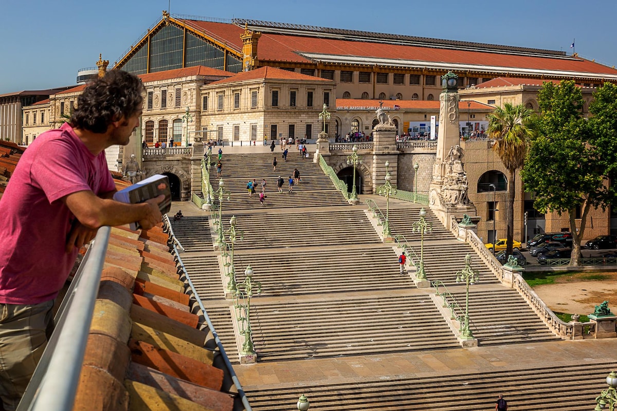 A view of the monumental staircase leading to the Saint-Charles railway station in Marseille. The staircase features multiple steps adorned with decorative posts, with pedestrians descending and ascending. Lush palm trees line the area, complementing the architecture of the station in the background.
