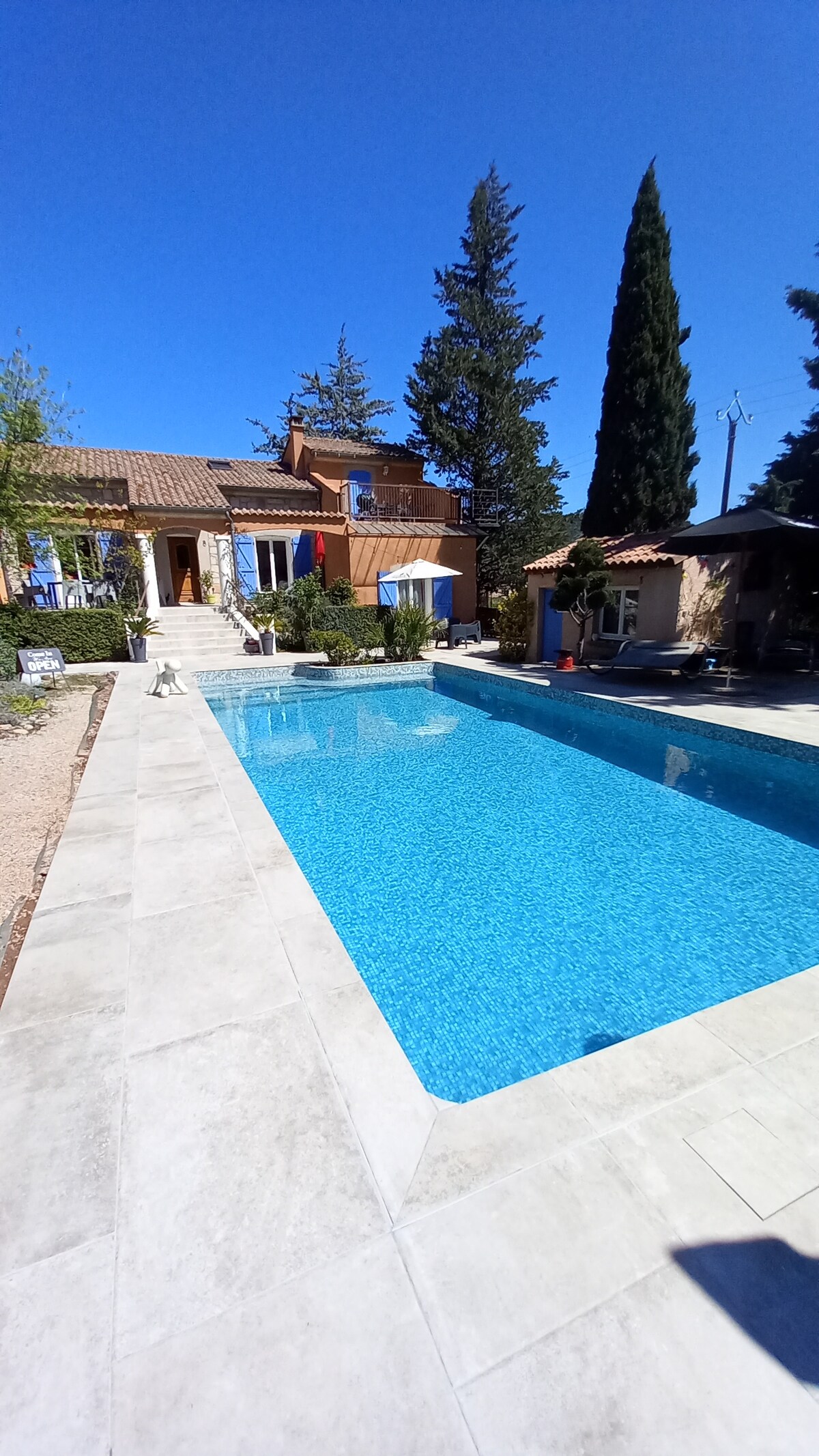 A clear blue pool reflects the sunny sky, surrounded by neat landscaping and trees. The property features a terracotta-colored building with a charming entrance. Lounge chairs are positioned beside the pool area, offering a serene outdoor space.
